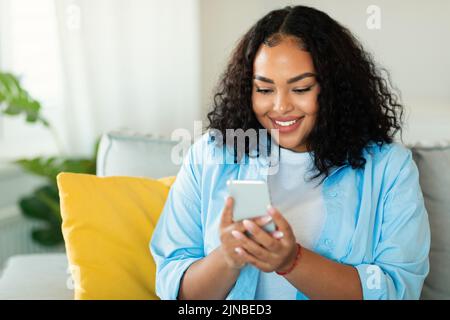 Allegra donna afroamericana che usa lo smartphone seduto a casa Foto Stock