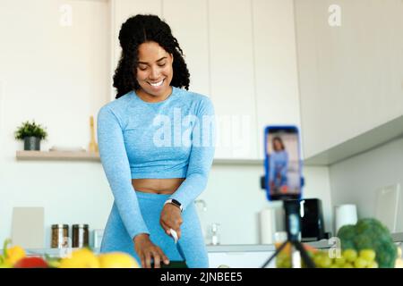 Donna afro-americana felice in sportswear registrazione video su smartphone fotocamera, preparare insalata fresca in cucina Foto Stock