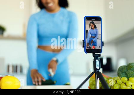 Donna afro-americana snella in abbigliamento sportivo preparazione insalata fresca e riprese video sulla fotocamera dello smartphone in cucina Foto Stock