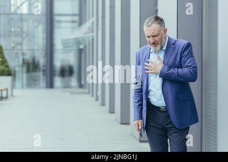 Il capo del banchiere grigio-haired anziano malato fuori dell'edificio dell'ufficio, banchiere ha dolore toracico severo, dolori di cuore Foto Stock