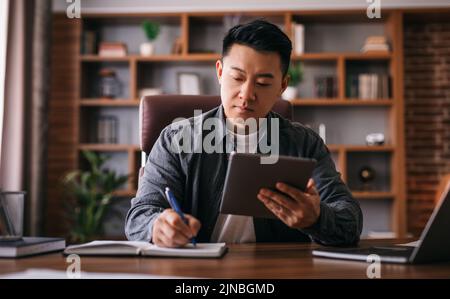 Occupato uomo cinese maturo al tavolo con notebook utilizza un tablet per lavorare in casa ufficio Foto Stock