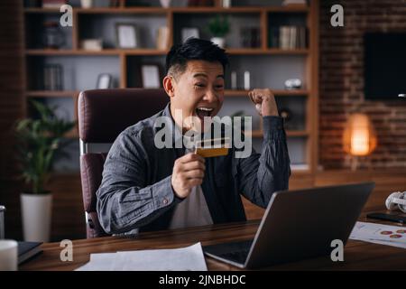 Felice uomo asiatico adulto con bocca aperta, urla, tenere la carta di credito, guarda il computer portatile e fa il gesto della vittoria Foto Stock
