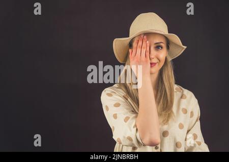 Giovane donna bionda bianca che indossa una camicia a pois di lino e un cappello di paglia che copre un occhio con la mano che guarda nella macchina fotografica. Ripresa da studio con sfondo scuro. Foto di alta qualità Foto Stock