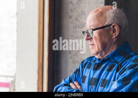 Uomo anziano in pensione calvo con le braccia incrociate, indossando i suoi occhiali e guardando pensieroso fuori dalla finestra. Foto di alta qualità Foto Stock