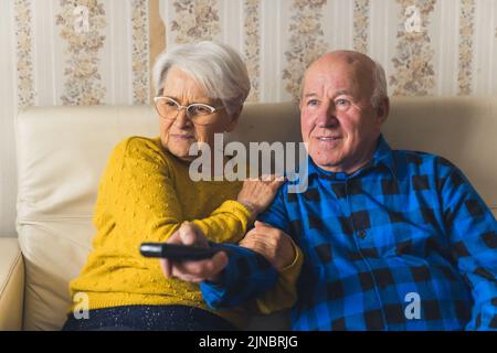 Coppia moderna anziana che guarda attivamente un film spaventoso o notizie su una tv, un uomo che tiene un telecomando. Foto di alta qualità Foto Stock