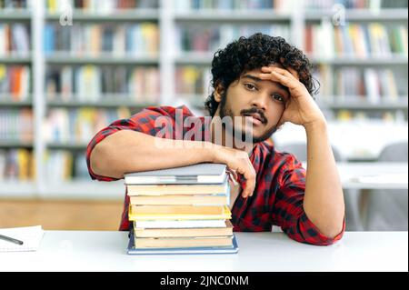 Stanco esausto ricci capelli indiano o arabo ragazzo, razza mista maschio, studente universitario, si siede su una scrivania con libri, in biblioteca sullo sfondo di librerie, guarda la macchina fotografica Foto Stock