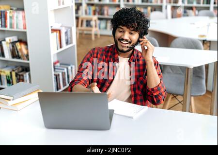 Gioioso moderno intelligente arabo o indiano studente maschio, con cuffie, si siede al tavolo con il computer portatile in biblioteca universitaria, parlando tramite videoconferenza, ascolta video corso on-line, discutere nuove informazioni Foto Stock