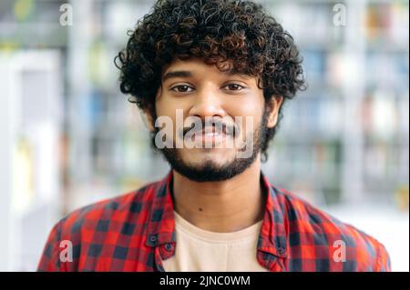 Foto ravvicinata di un bel ragazzo indiano o arabo dai capelli ricci positivi, in camicia casual, manager, designer, studente o freelance, si trova all'interno, guarda la fotocamera, sorridente amichevole Foto Stock