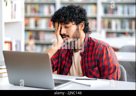 Studente stanco di nazionalità araba o indiana addormentato ad una scrivania in biblioteca, in abiti eleganti, stanco di prepararsi per l'esame, leggendo libri, si sente esausto, chiuso gli occhi, ha bisogno di riposo Foto Stock