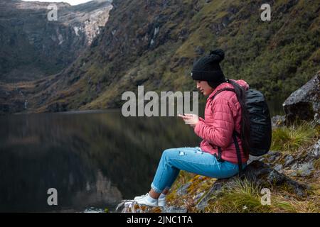 Una ragazza caucasica in una giacca rosa con uno zaino seduto su una roccia sul lago e guardando il suo telefono in montagna