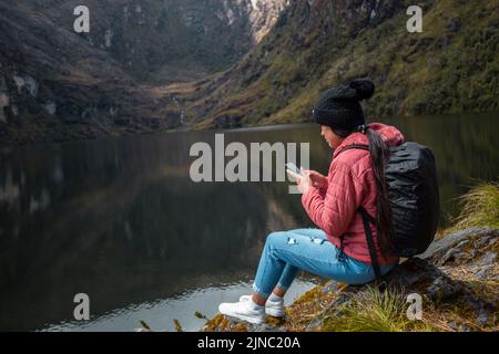 Una ragazza caucasica in una giacca rosa con uno zaino seduto su una roccia sul lago e guardando il suo telefono in montagna