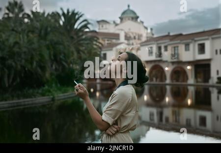 La classe è senza tempo: Una giovane donna attraente che fuma una sigaretta accanto a un lago in un ambiente urbano. Foto Stock