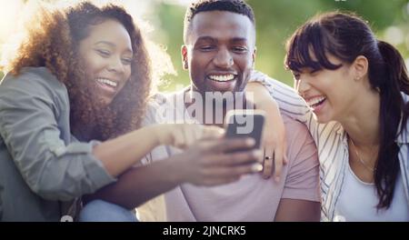 Guardate cosa tutti stanno facendo pazzi online. Un allegro gruppo di amici che usano un cellulare insieme mentre si rilassano all'aperto. Foto Stock