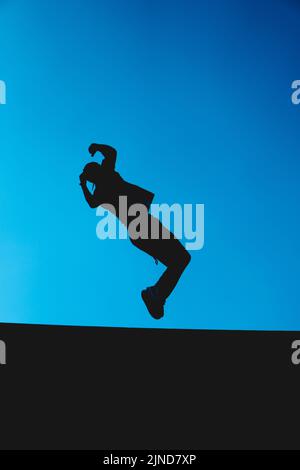 Silhouette di un uomo che fa parkour con il cielo blu sullo sfondo. Somersault estremo. Ombra di un atleta che salta indietro. Foto Stock