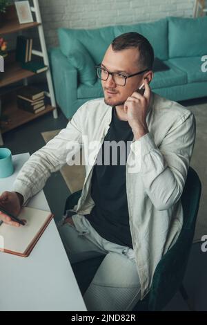 Un uomo d'affari concentrato che tiene il telefono nelle mani, scrivendo a mano informazioni importanti in ufficio. Uomo che utilizza un telefono cellulare, comunica con i partner. Foto Stock
