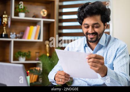 Felice giovane uomo sorridente che legge l'offerta o la lettera promozionale mentre si siede all'ufficio - concetto di realizzazione, approvazione di prestito e promozione di lavoro Foto Stock