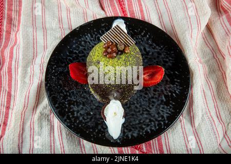 Torta dolce singola. Torta al cioccolato smaltata, un pezzo, su piatto nero con torta al pistacchio. Viene servito su un piatto con gelato e fragole. Foto Stock