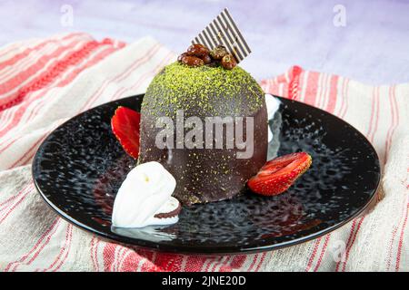 Torta dolce singola. Torta al cioccolato smaltata, un pezzo, su piatto nero con torta al pistacchio. Viene servito su un piatto con gelato e fragole. Foto Stock