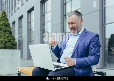 Un uomo d'affari dai capelli grigi di successo che lavora su un computer portatile per celebrare la vittoria trionfa un uomo sovrappeso seduto su una panca all'esterno dell'edificio dell'ufficio Foto Stock