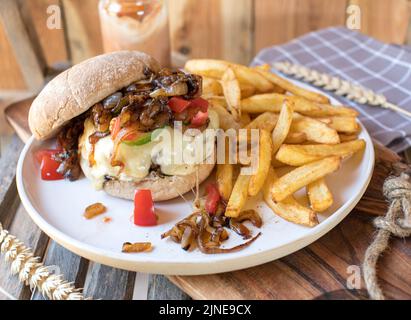 Hamburger fatto in casa con cipolle arrostite, pomodori e cetriolo. Servito con patatine fritte su sfondo di legno Foto Stock