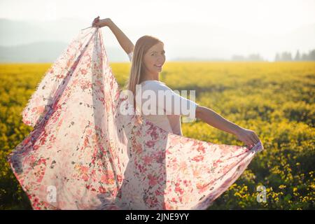 Giovane donna capelli lunghi che balla in campo fiori gialli, sciarpa che sta tenendo in movimento dal vento, pomeriggio luce del sole sfondo Foto Stock