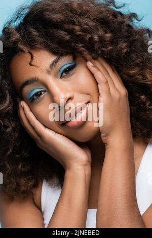 ritratto di donna afroamericana con trucco che tocca il viso e sorridente alla macchina fotografica isolato su blu Foto Stock