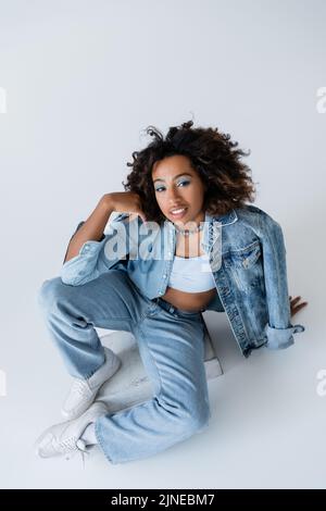 vista ad alto angolo di una donna afro-americana in elegante abbigliamento denim seduto su sfondo grigio Foto Stock