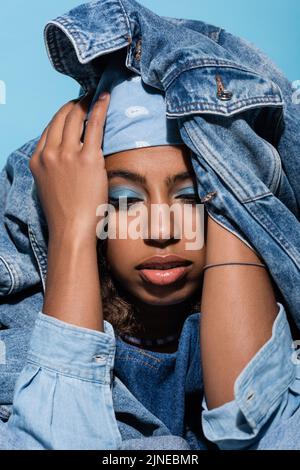 donna afroamericana con capo coprente trucco con giacca in denim isolato su blu Foto Stock