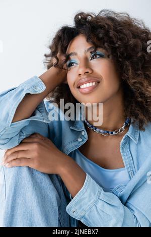 gioiosa donna afroamericana in perline e camicia blu in denim guardando lontano isolato sul grigio Foto Stock