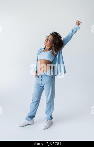 intera lunghezza di elegante donna afroamericana con mano rialzata in piedi su sfondo grigio Foto Stock