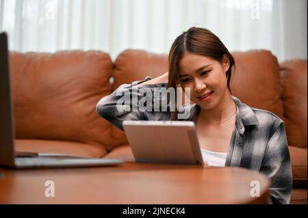 Bella giovane donna asiatica utilizza il touchpad digitale del tablet per chattare con i suoi amici online mentre si rilassa nel suo salotto. Foto Stock