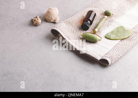 Olio per il viso, utensili da massaggio gua sha e asciugamano di cotone su sfondo grigio in pietra con spazio copia. Trattamento della pelle, concetto di spa casalinga. Foto Stock