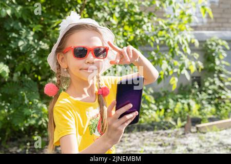 Ritratto di bambina che prende un selfie da smartphone nel parco estivo. Una ragazza allegra in un vestito giallo, cappello bianco e occhiali da sole prende un Foto Stock