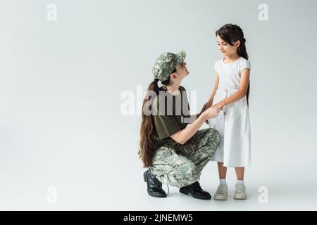 Donna sorridente in uniforme militare tenendo le mani di figlia su sfondo grigio, immagine stock Foto Stock