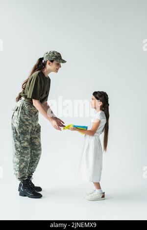 Vista laterale del bambino che tiene bandiera ucraina vicino alla madre in uniforme militare su sfondo grigio, immagine stock Foto Stock