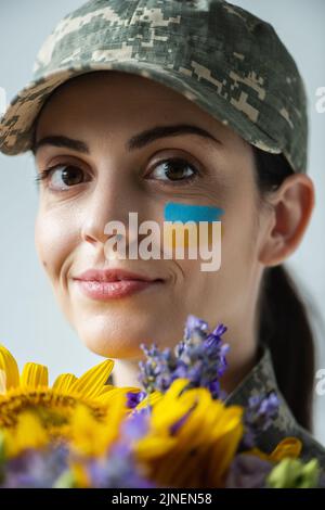 primo piano ritratto di felice ucraino servicewoman vicino a fiori blu e giallo isolato su grigio, immagine stock Foto Stock