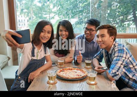 Giovane donna vietnamita che prende selfie con i suoi amici e grande pizza Foto Stock