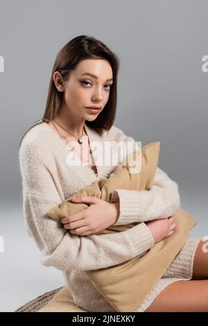 giovane donna in maglione e collane dorate che abbracciano il cuscino su sfondo grigio, immagine stock Foto Stock