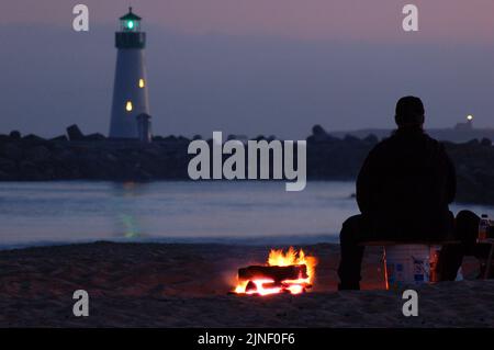 Un uomo adulto siede accanto a un falò davanti a un faro sulla spiaggia del Twin Lakes state Park, Santa Cruz, California Foto Stock