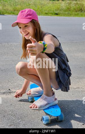 Ritratto di una ragazza caucasica sorridente con capelli lunghi in un cappellino rosa da baseball, seduta su uno skateboard su una strada asfaltata in condizioni di sole. Un bambino cavalca Foto Stock