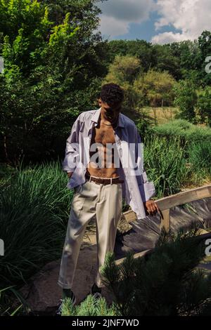 uomo afroamericano in camicia blu e pantaloni bianchi in piedi al lago nel parco, immagine stock Foto Stock