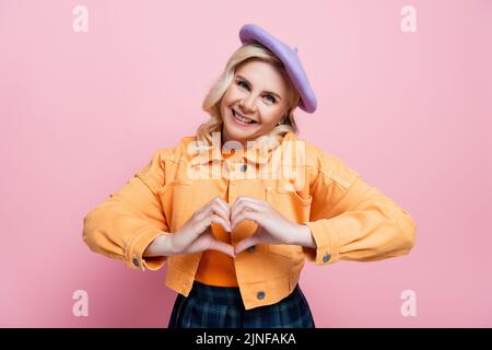 Donna bionda sorridente in beretta e giacca mostrando segno d'amore isolato su rosa, immagine stock Foto Stock