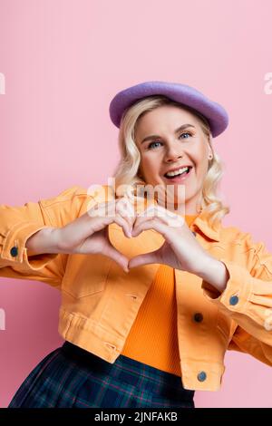 Donna bionda positiva in beretta che mostra segno di cuore isolato su rosa, immagine stock Foto Stock