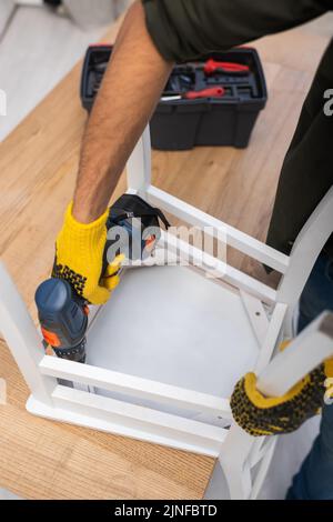 Vista ritagliata di un uomo nei guanti che ripara la sedia con un cacciavite elettrico vicino alla cassetta degli attrezzi sfocata a casa, immagine stock Foto Stock