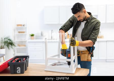 Uomo musulmano in guanti sedia di fissaggio con trapano elettrico vicino cassetta degli attrezzi sul tavolo a casa, immagine stock Foto Stock