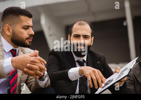 Un gruppo di uomini d'affari si riunisce all'aperto. I dipendenti di sesso maschile in tute comunicano all'aperto. I partner commerciali discutono e discutono. Relazioni e. Foto Stock