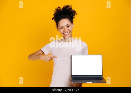 Felice ragazza afro-americana, tenendo un notebook aperto con schermo bianco in mano, punta il dito su di esso, si trova su sfondo arancione isolato, guardando la fotocamera, sorriso amichevole. Concetto di mock-up Foto Stock