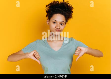 Negativo pessimista giovane afroamericana con capelli ricci, vestita in t-shirt di base, con un gesto verso il basso pollici, guarda la macchina fotografica, in piedi su sfondo isolato arancione, triste espressione facciale Foto Stock