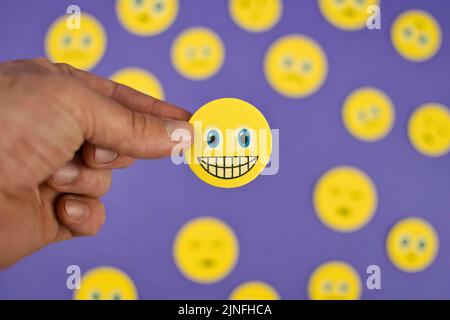 Primo piano scatto di mano che tiene giallo emotoji sorridente faccia tra emotis triste su sfondo viola Foto Stock
