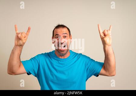 Gesti dei fan della roccia. Uomo caucasico barbuto in t-shirt blu che fa il simbolo del rock con le mani in su mentre si trova su sfondo beige. Linguaggio del corpo Foto Stock